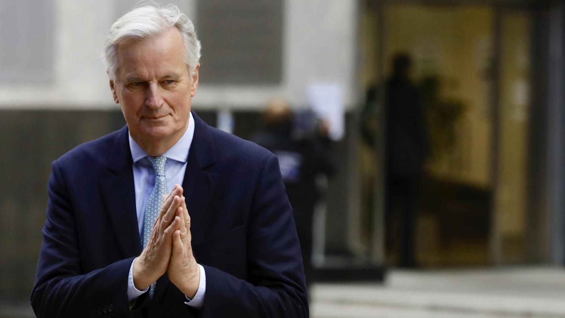 Barnier steht vor einem Eingang und hält die Hände wie beim Beten zusammen.