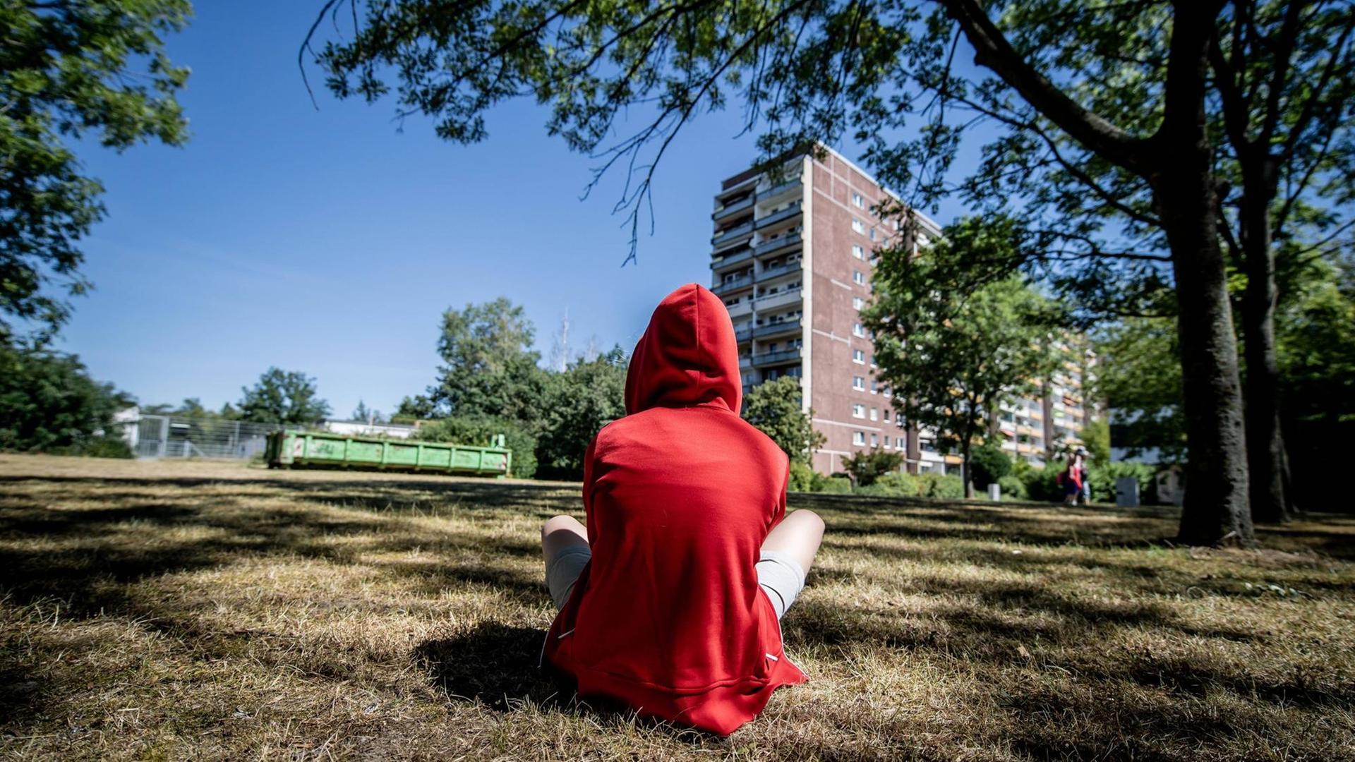 Ein jugendliches Kind sitzt in einem roten Kapuzenpullover mit dem Rücken zum Betrachter auf einer Wiese. Im Hintergrund ist ein Plattenbau zu sehen. Ein jugendliches Kind sitzt in einem roten Kapuzenpullover mit dem Rücken zum Betrachter auf einer Wiese. Im Hintergrund ist ein Plattenbau zu sehen.