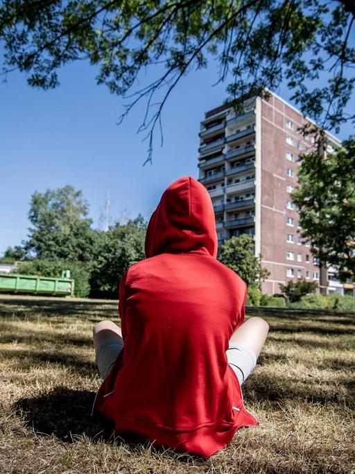 Ein jugendliches Kind sitzt in einem roten Kapuzenpullover mit dem Rücken zum Betrachter auf einer Wiese. Im Hintergrund ist ein Plattenbau zu sehen.
