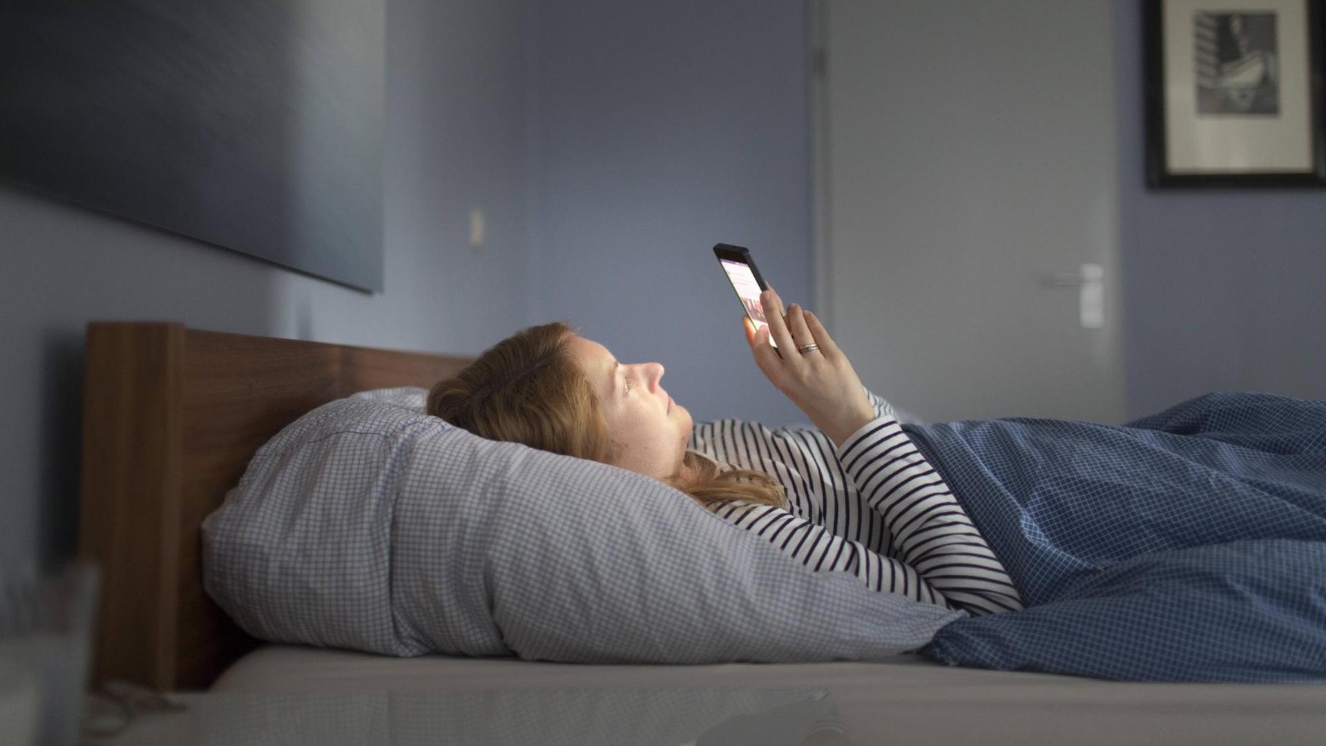 Frau liegt im Bett in einem abgedunkelten Raum und blickt auf ihr Smartphone Frau liegt im Bett in einem abgedunkelten Raum und blickt auf ihr Smartphone