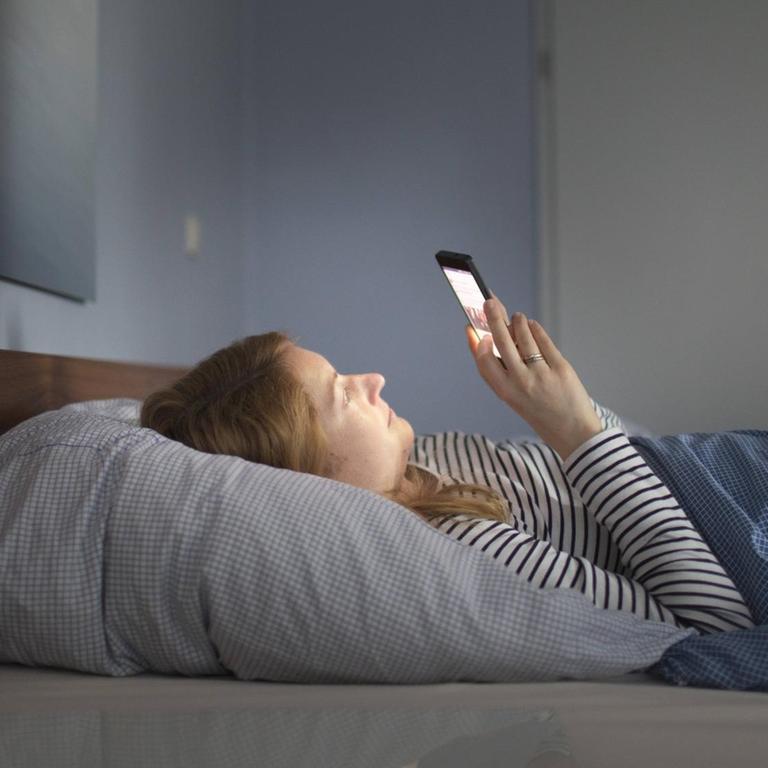 Frau liegt im Bett in einem abgedunkelten Raum und blickt auf ihr Smartphone