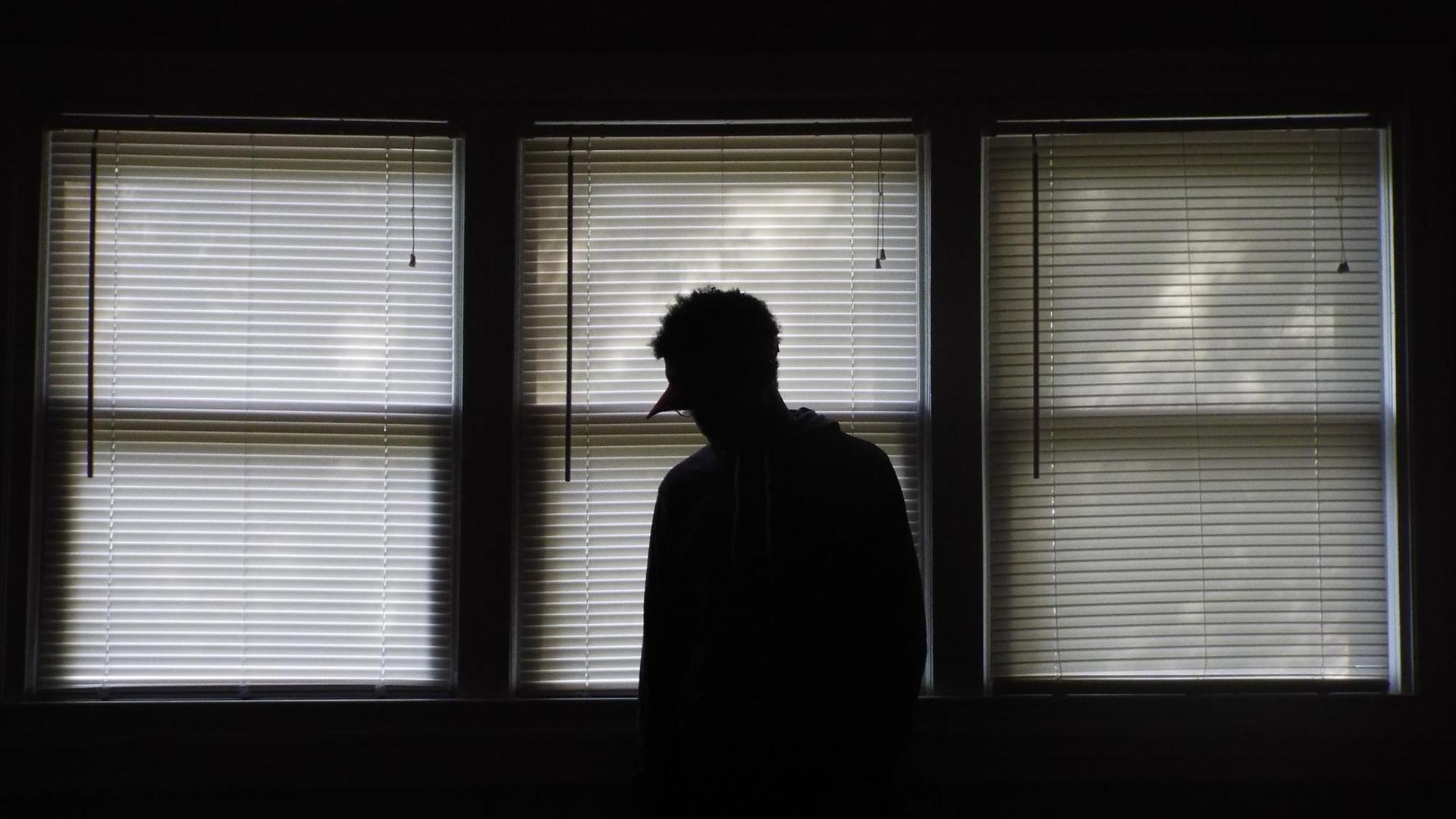Die Silhouette einer Person vor drei Fenstern, an denen die Rollos runtergelassen sind. Die Silhouette einer Person vor drei Fenstern, an denen die Rollos runtergelassen sind.