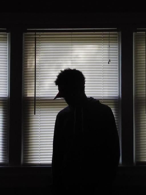 Die Silhouette einer Person vor drei Fenstern, an denen die Rollos runtergelassen sind.