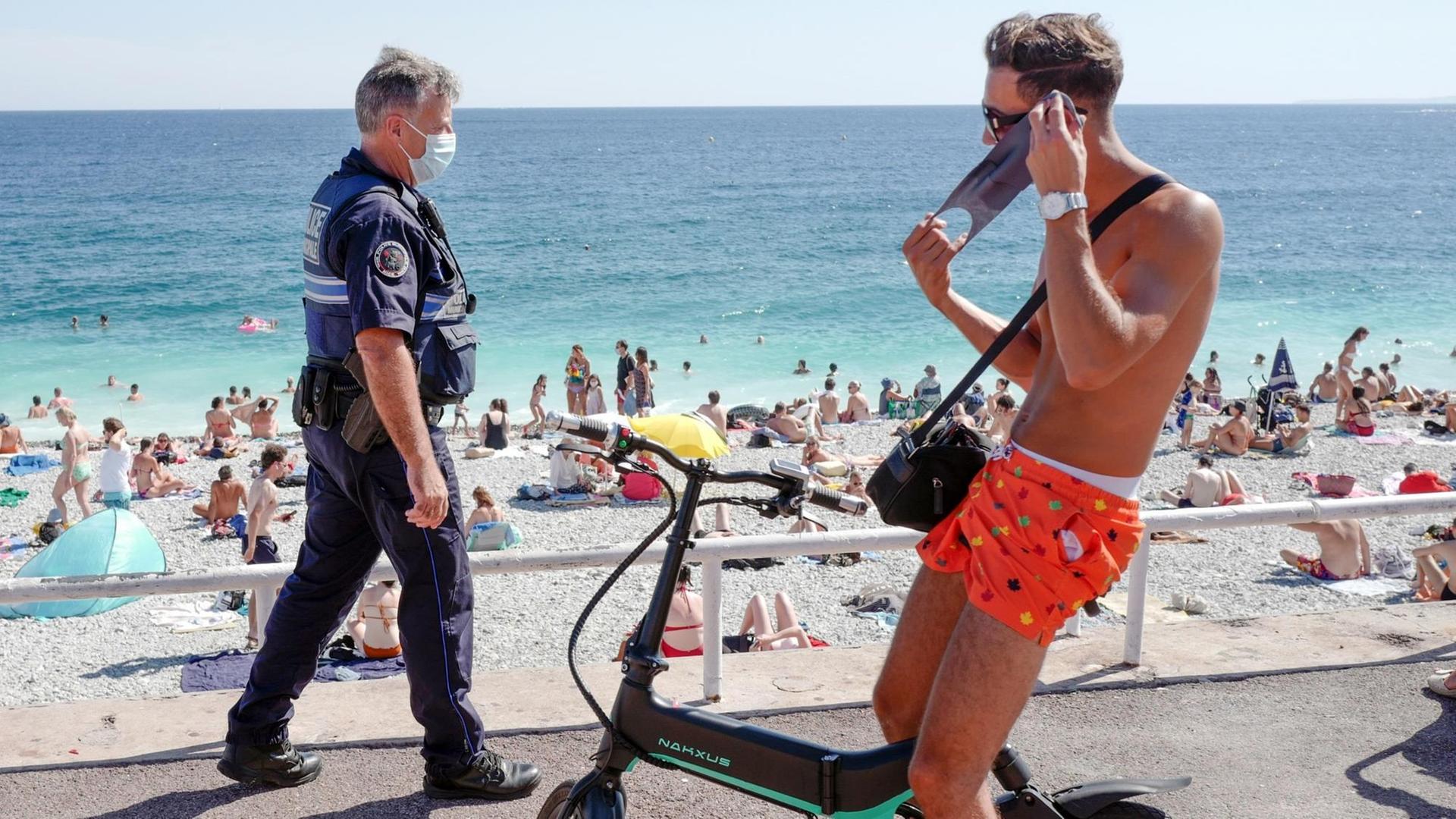 Ein Polizist mit Maske patroulliert auf der Promenade. Im Vordergrund legt ein junger Radfahrer in Badeshorts einen Mundschutz an.