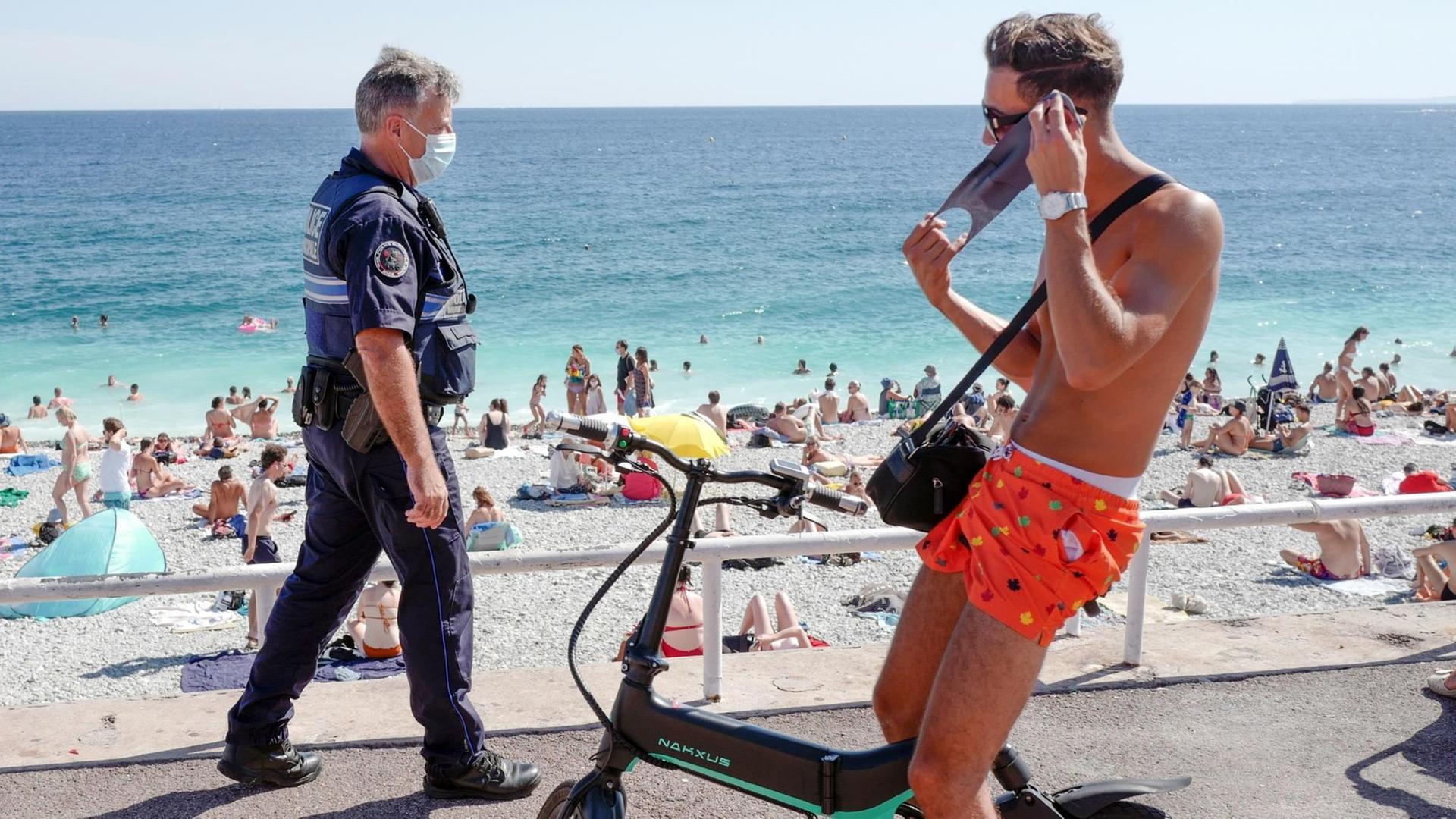 Ein Polizist mit Maske patroulliert auf der Promenade. Im Vordergrund legt ein junger Radfahrer in Badeshorts einen Mundschutz an. Ein Polizist mit Maske patroulliert auf der Promenade. Im Vordergrund legt ein junger Radfahrer in Badeshorts einen Mundschutz an.
