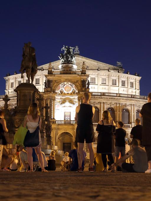 Menschen stehen vor der Semperoper in Dresden und lauschen dem Gesang eines Tenors und einer Sopranistin, die in einer warmen Sommernacht Opernarien darbieten