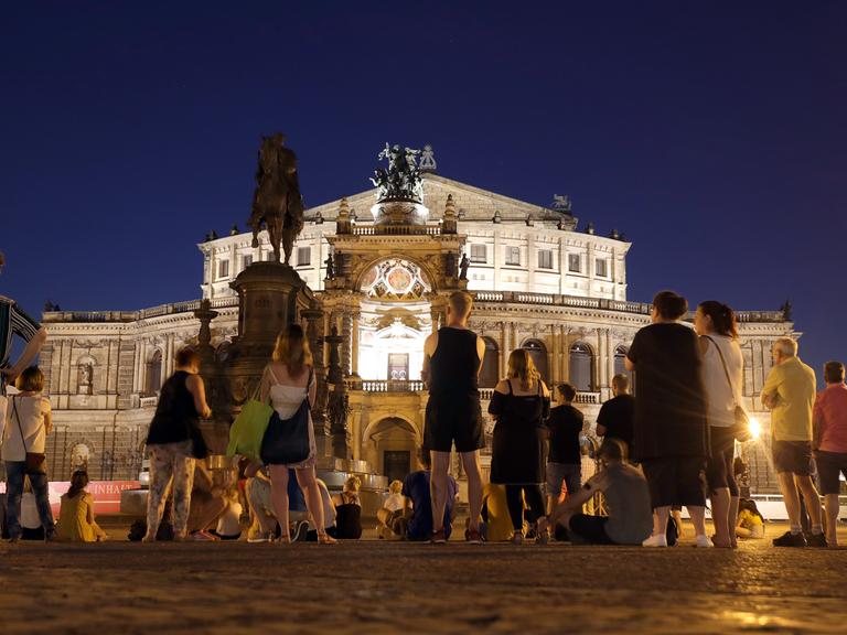 Menschen stehen vor der Semperoper in Dresden und lauschen dem Gesang eines Tenors und einer Sopranistin, die in einer warmen Sommernacht Opernarien darbieten