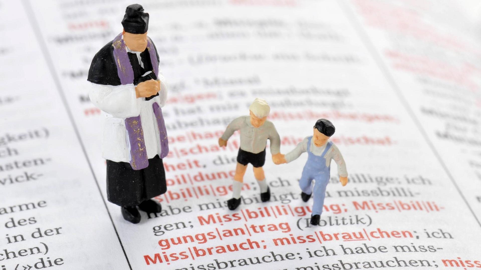 Modelleisenbahnfiguren eines Priesters und zweier Kinder auf einer aufgeschlagenen Wörterbuchseite vor dem Wort "Missbrauch"