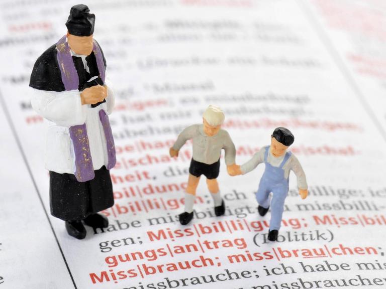 Modelleisenbahnfiguren eines Priesters und zweier Kinder auf einer aufgeschlagenen Wörterbuchseite vor dem Wort "Missbrauch"