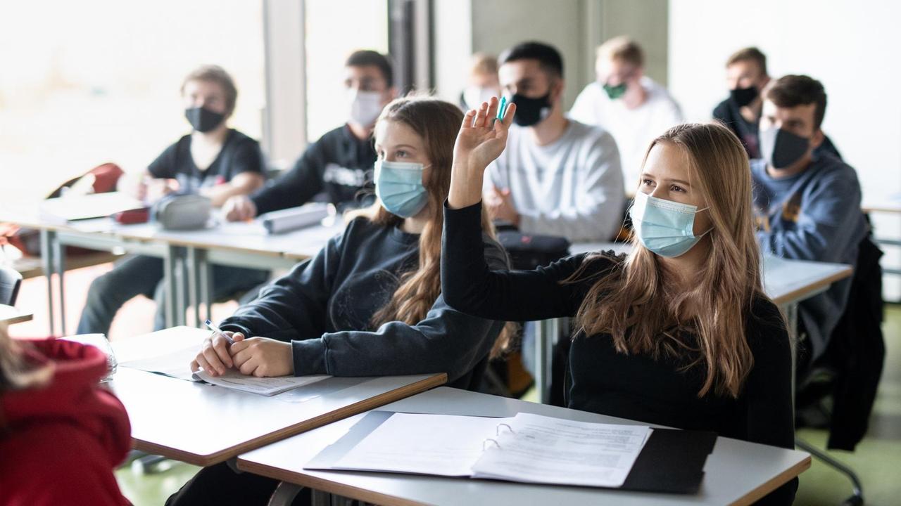 Schülerinnen und Schüler nehmen mit Mund- und Nasenschutz am Unterricht teil, eine Schülerin meldet sich. Schülerinnen und Schüler nehmen mit Mund- und Nasenschutz am Unterricht teil, eine Schülerin meldet sich.