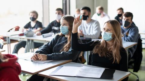 Schülerinnen und Schüler nehmen mit Mund- und Nasenschutz am Unterricht teil, eine Schülerin meldet sich.