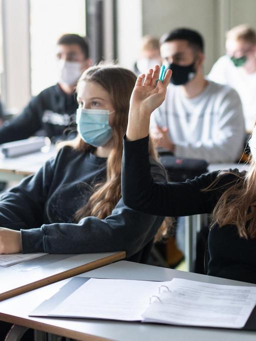 Schülerinnen und Schüler nehmen mit Mund- und Nasenschutz am Unterricht teil, eine Schülerin meldet sich.