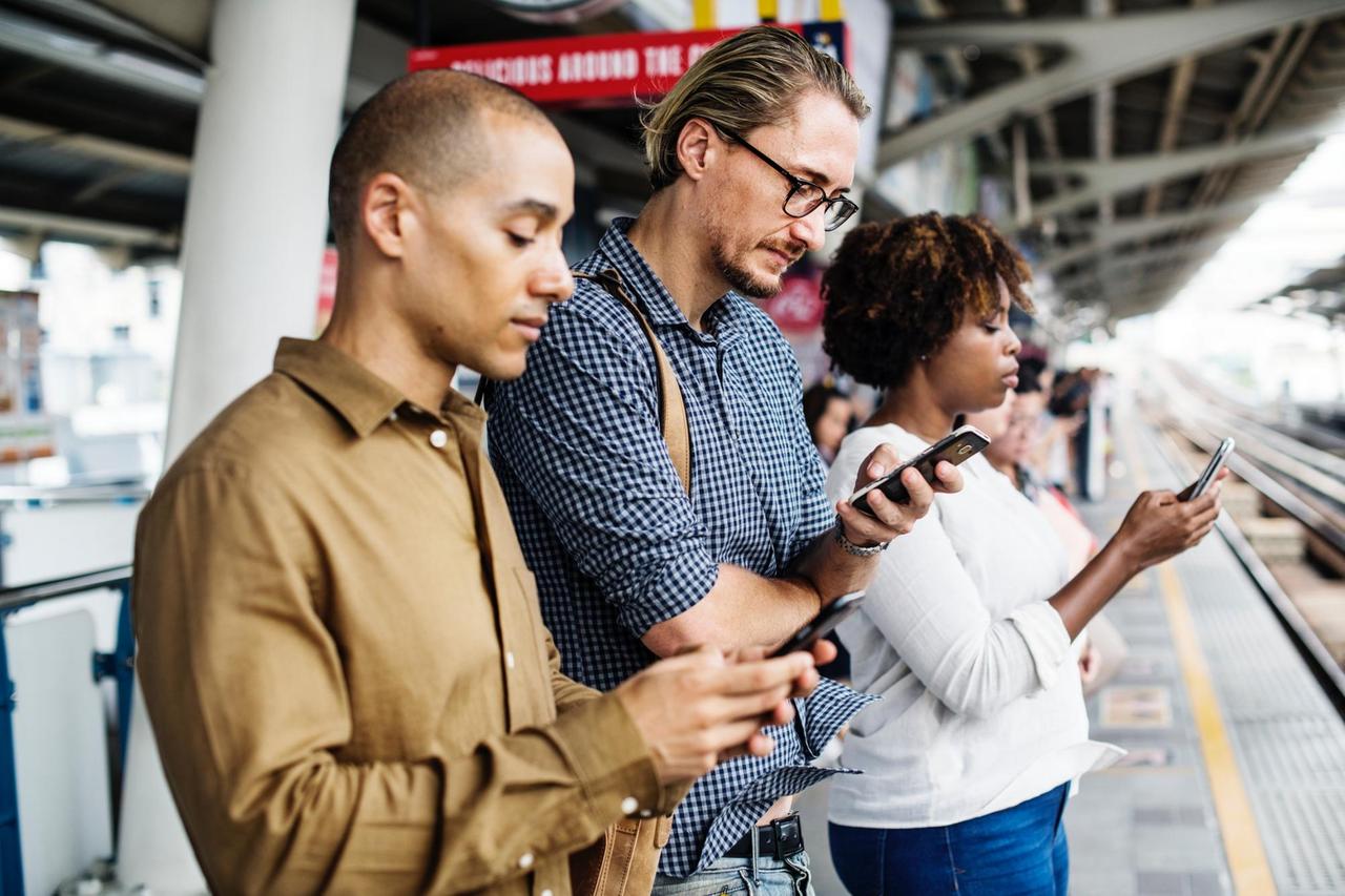 Drei Menschen stehen nebeneinander auf einem Bahnsteig und gucken auf ihre Smartphones Drei Menschen stehen nebeneinander auf einem Bahnsteig und gucken auf ihre Smartphones