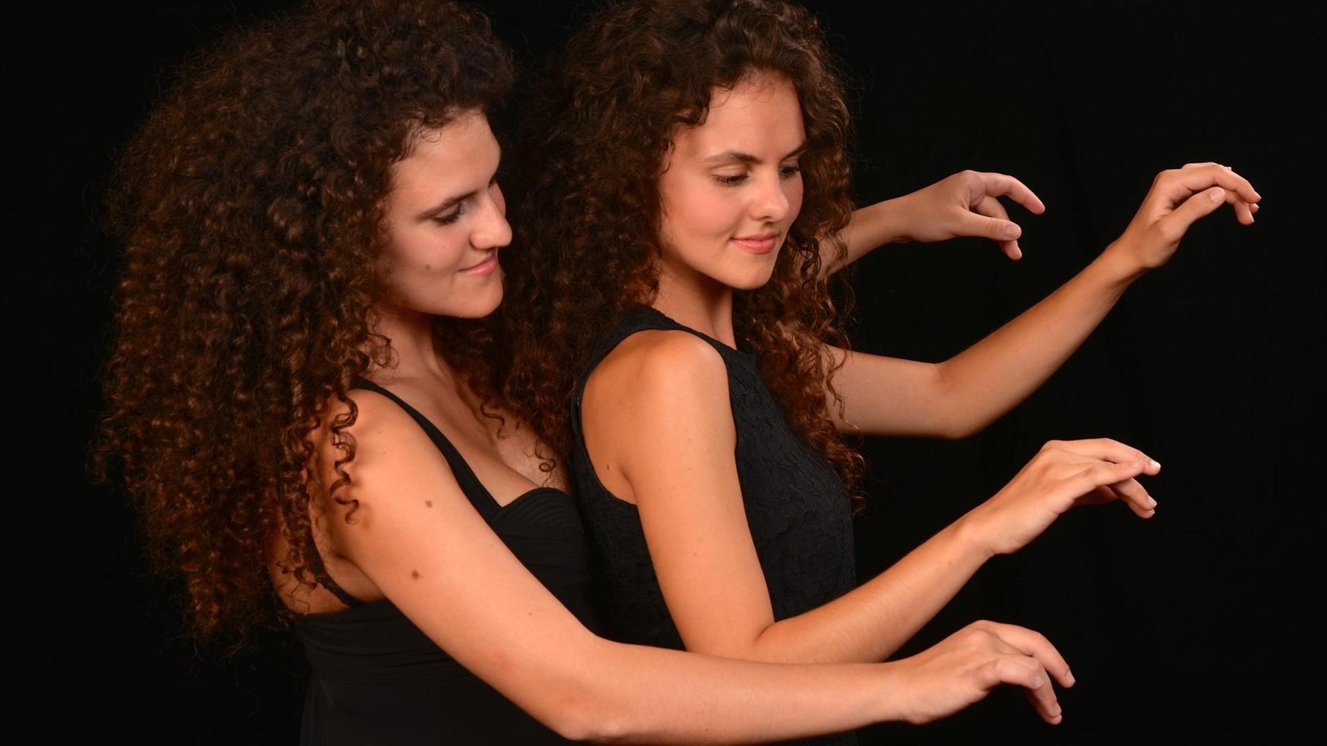 Zwei junge Frauen in schwarzen Kleidern sitzen hintereinander an einem imgaginierten Flügel und spielen.