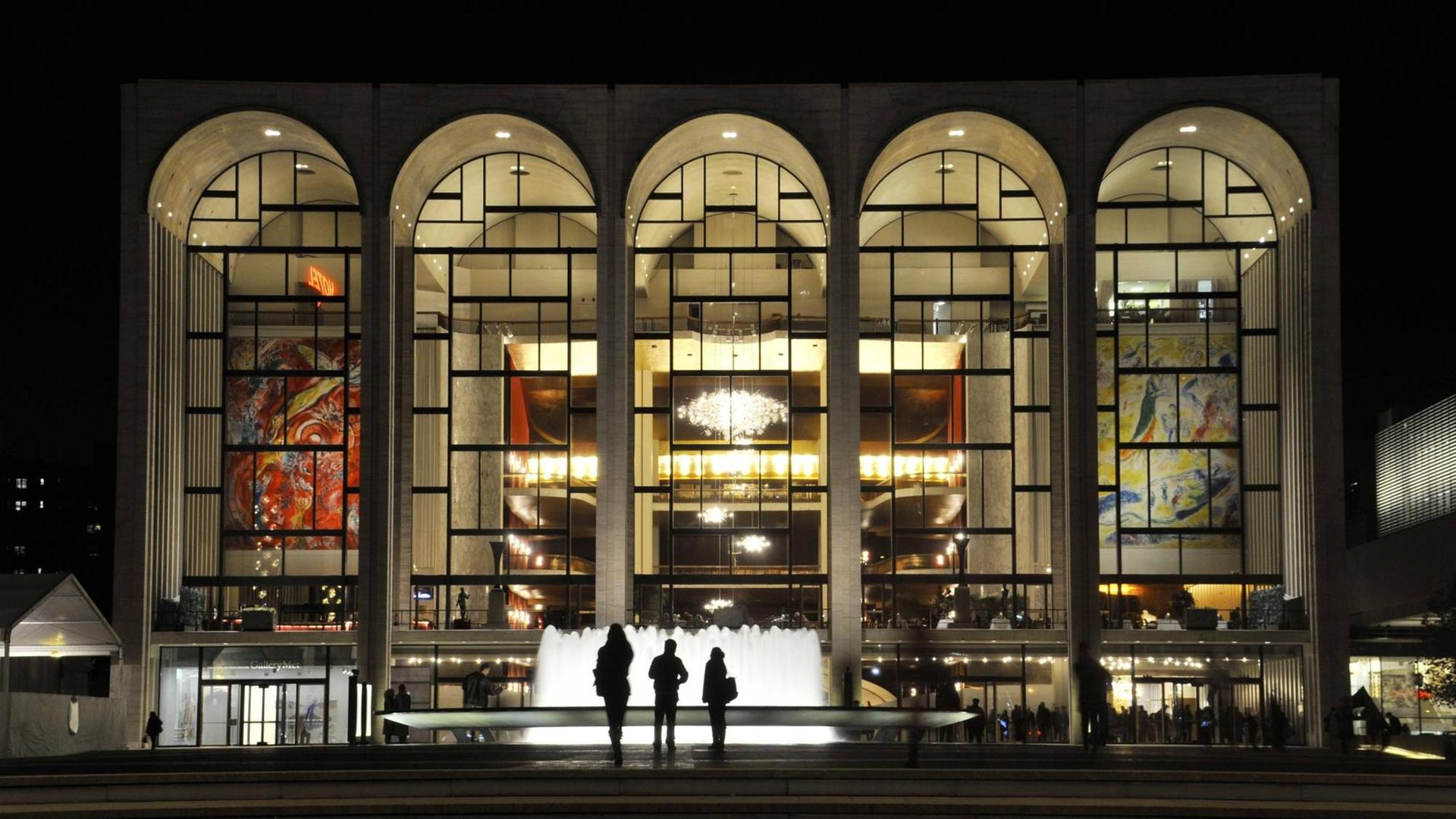Menschen stehen im Dunklen vor einem Springbrunnen und blicken in die fünf hell erleuchteten, großen bogenförmigen Fenster des Metropolitan Opera House in New York City. Menschen stehen im Dunklen vor einem Springbrunnen und blicken in die fünf hell erleuchteten, großen bogenförmigen Fenster des Metropolitan Opera House in New York City.