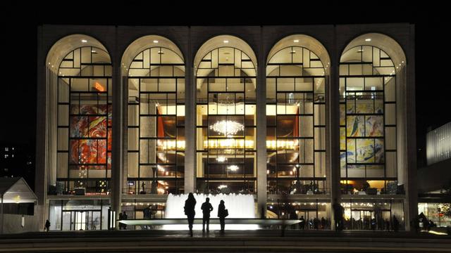 Menschen stehen im Dunklen vor einem Springbrunnen und blicken in die fünf hell erleuchteten, großen bogenförmigen Fenster des Metropolitan Opera House in New York City.
