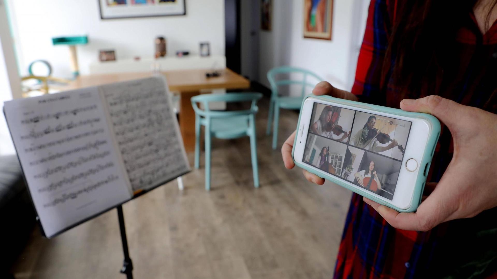 In einem hellen Zimmer mit Esstisch und Stühlen steht ein Nontenständer, neben dem jemand ein Handy präsentiert, auf dem vier Musiker zu sehen sind, die gemeinsam mit Streichinstrumenten bei einer Schalte spielen. In einem hellen Zimmer mit Esstisch und Stühlen steht ein Nontenständer, neben dem jemand ein Handy präsentiert, auf dem vier Musiker zu sehen sind, die gemeinsam mit Streichinstrumenten bei einer Schalte spielen.