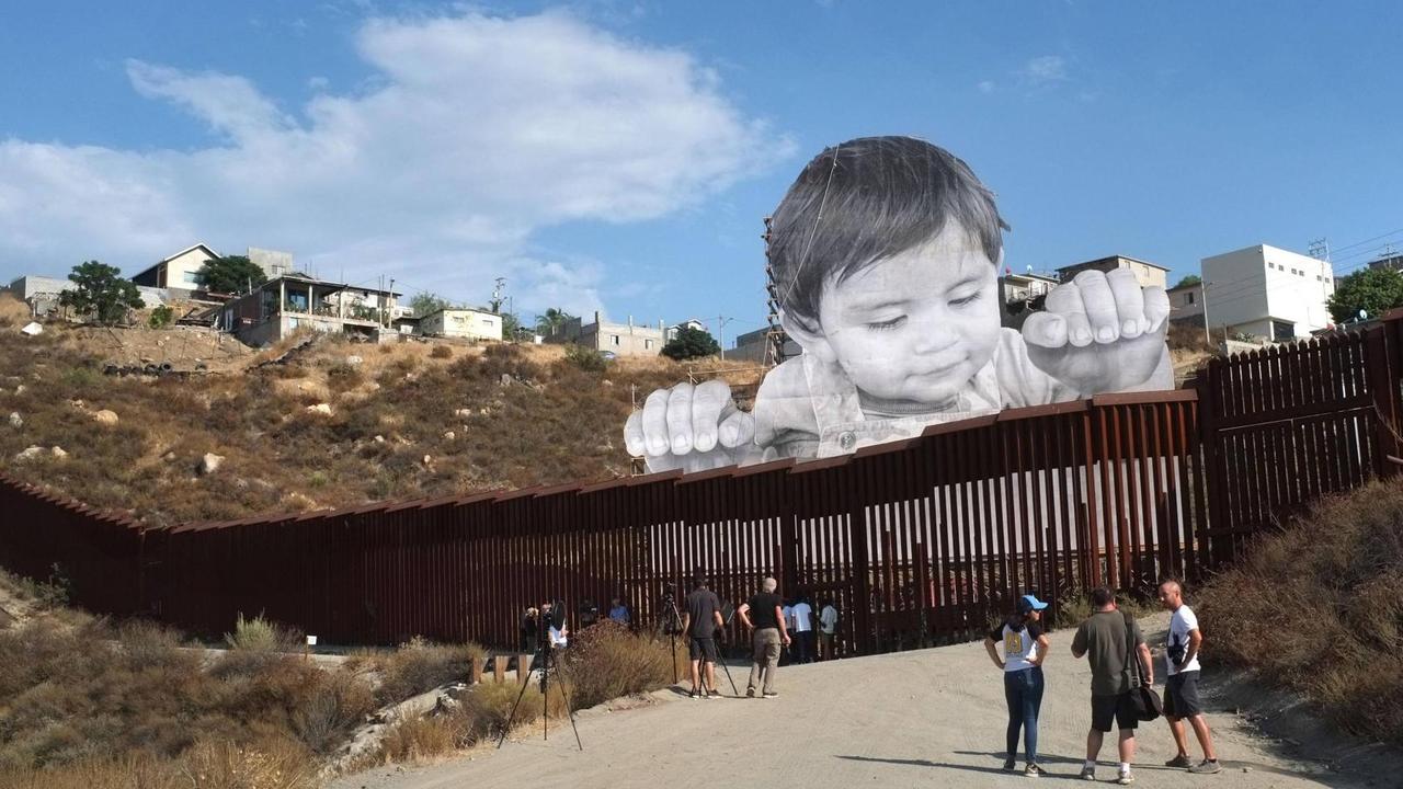 Die Installation des französischen Streetart-Künstlers JR an der Grenzmauer zwischen den USA und Mexiko: Leute stehen vor der Mauer und sehen sich das überlebensgroße Foto eines Kindes an, das über die Mauer schaut. Die Installation des französischen Streetart-Künstlers JR an der Grenzmauer zwischen den USA und Mexiko: Leute stehen vor der Mauer und sehen sich das überlebensgroße Foto eines Kindes an, das über die Mauer schaut.