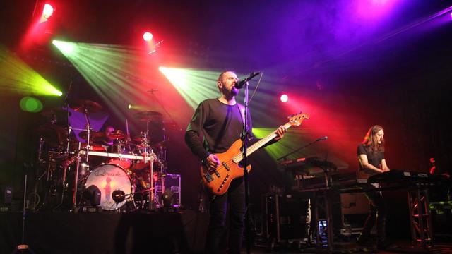 Drei Männer an Schlagzeug, Bass und Keyboards musizieren auf einer bunt beleuchteten Bühne.