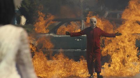 Im Still aus "Wir" steht ein Maskierter mit ausgebreiteten Armen vor einem brennenden Auto.