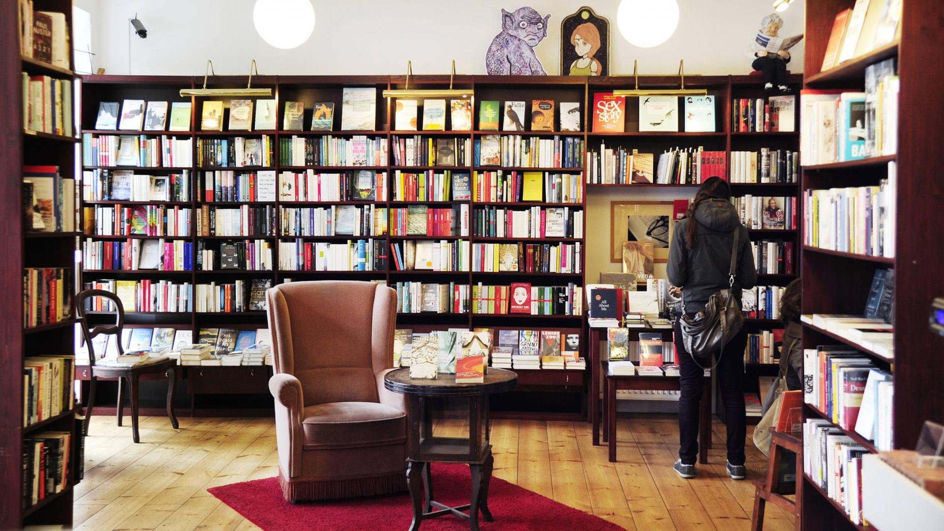 Eine Buchhandlung mit vielen Regalen. Ein Mann steht vor einem Regal und liest. In der Mitte des Raums steht ein gemütlicher Ohrensessel.