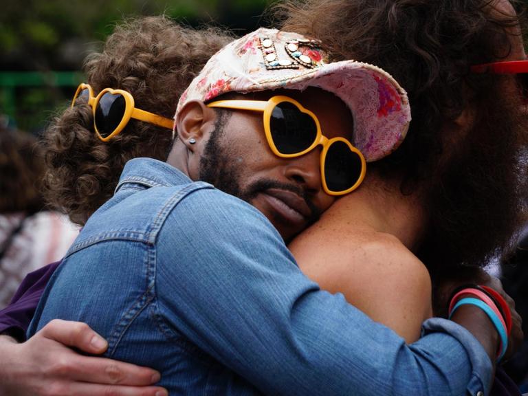 Ein Mann mit gelber Herzbrille umarmt einen anderen Mann von hinten und schmiegt sich an seinen Rücken, im Hintergrund sieht man noch mehr Menschen, alle tragen Plastikherzbrillen