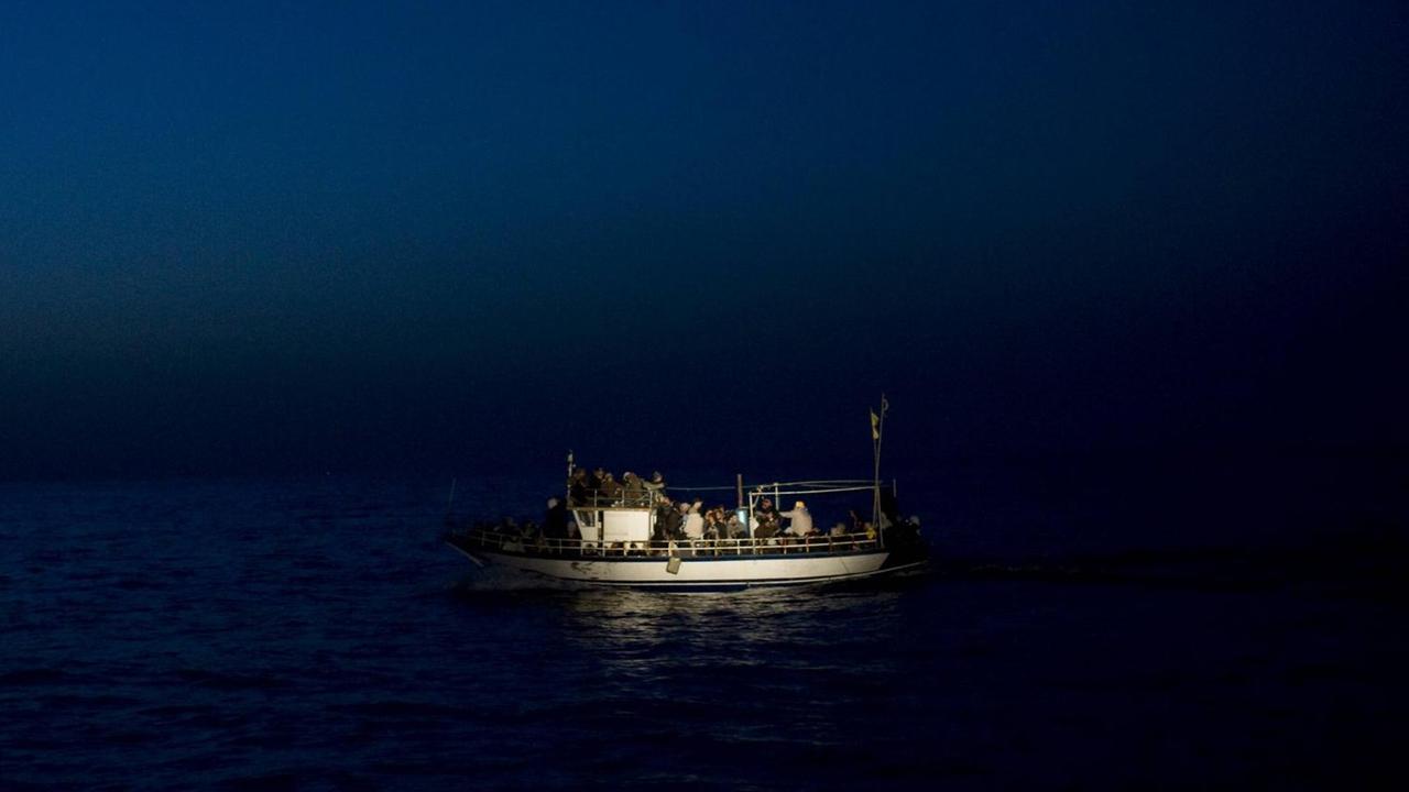 Ein kleines Boot mit vielen Menschen liegt auf dem Wasser. Es ist Nacht, das Boot wird angestrahlt. Ein kleines Boot mit vielen Menschen liegt auf dem Wasser. Es ist Nacht, das Boot wird angestrahlt.