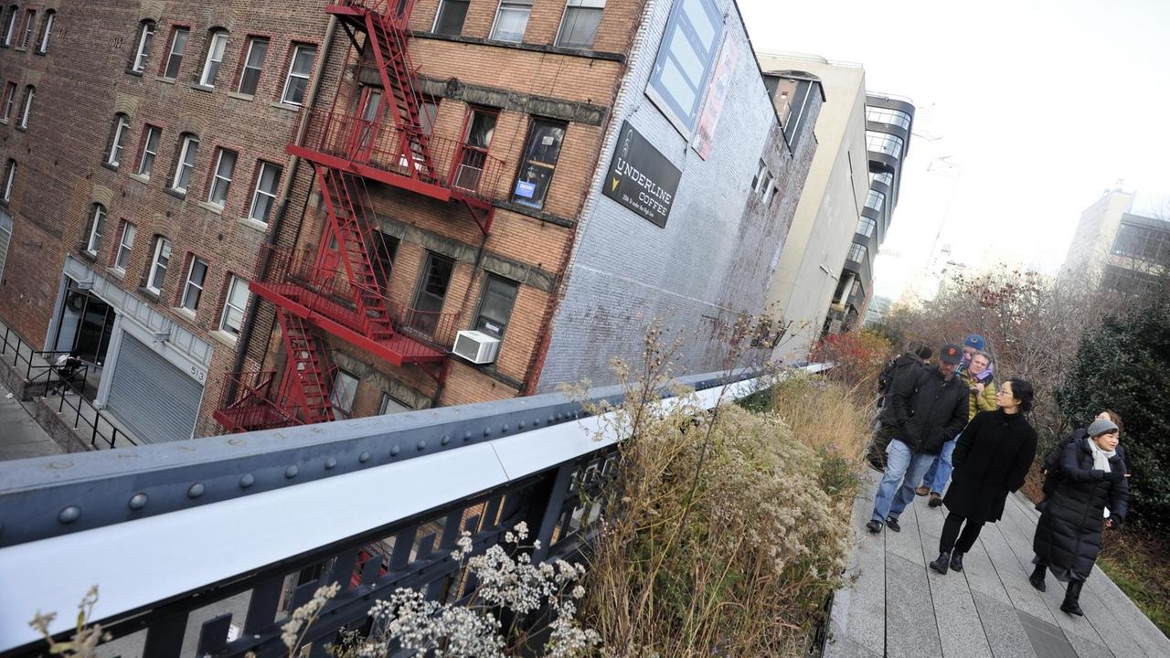 Männer und Frauen in warmer Kleidung spazieren über die "Chelsea Highline", einen begrünten Weg auf einer ehemaligen Hochbahntrasse, durch Manhattan, links von der Brüstung ist ein Backsteinhaus mit roten Feuertreppen aus Metall zu sehen. Männer und Frauen in warmer Kleidung spazieren über die "Chelsea Highline", einen begrünten Weg auf einer ehemaligen Hochbahntrasse, durch Manhattan, links von der Brüstung ist ein Backsteinhaus mit roten Feuertreppen aus Metall zu sehen.