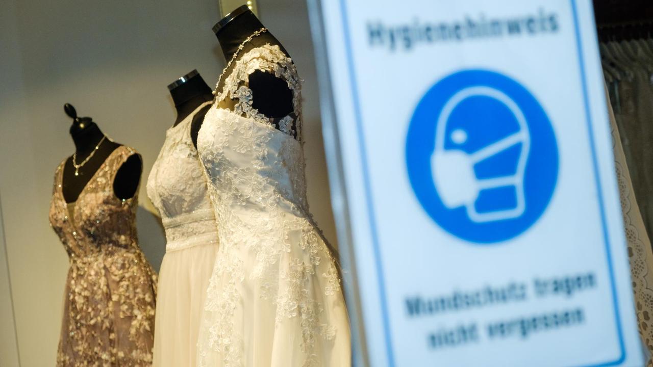 Brautkleider sind in einem Schaufenster eines Brautmoden-Geschäfts zu sehen. Im Vordergrund steht ein Schild, das auf die Maskenpflicht während der Corona-Pandemie verweist.