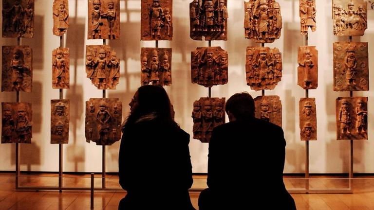 Zwei Menschen sitzen im Museum vor kunstvoll verzierten Steinplatten aus dem westafrikanischen Benin.