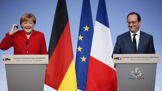 Sie sehen Bundeskanzlerin Merkel und Frankreichs Präsident Hollande, zwischen ihnen stehen Fahnen. Beide lachen.
