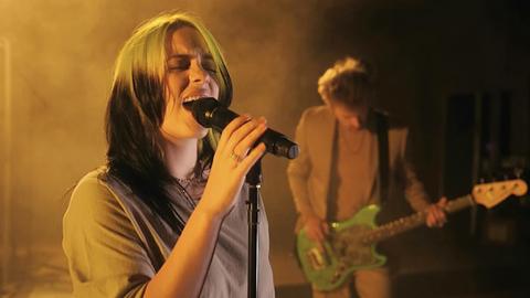 Die Singer-Songwriterin Billie Eilish steht am Mikrofon und singt mit geschlossenen Augen. Im Hintergrund ist gelblicher Rauch zu sehen sowie ein Musiker, der mit gesenktem Kopf Gitarre spielt.