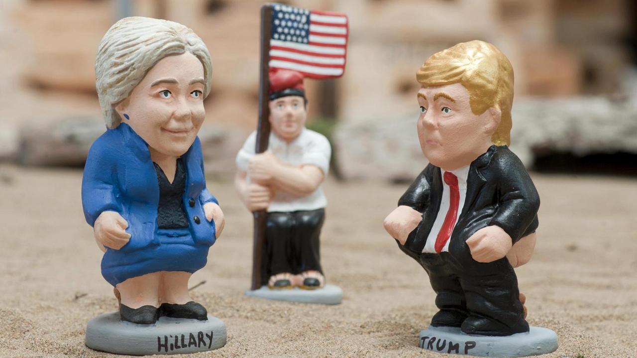 Zwei Spielzeug-Figuren, die Hillary Clinton und Donald Trump darstellen, im Hintergrund eine weitere Figur, die eine US-Flagge hält.
