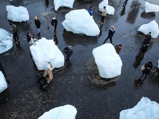 Auf einem Platz liegen große Eisblöcke. Zwischen den Eisblöcken laufen Menschen herum.