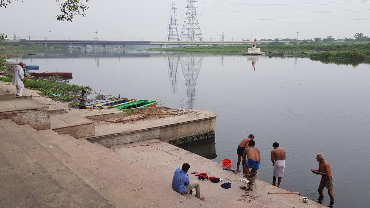 An den Treppen, die zum heiligen Fluss Yamuna in der indischen Hauptstadt Delhi hinunterführen, stehen Menschen in Badehosen oder mit um die Hüfte geschlungenen Tüchern bis zu den Knöcheln im Wasser, um sich damit zu benetzen.