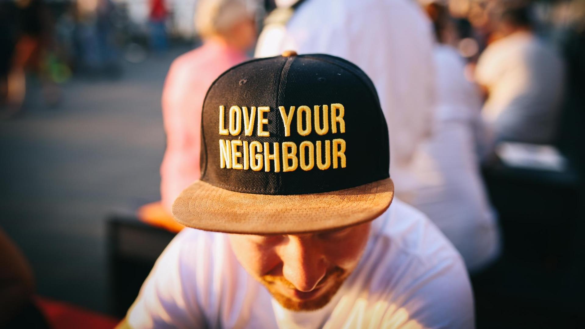 Ein junger Mann mit einer Baseball-Kappe auf der "Love you Neighbour" in knallgelben Lettern steh, sitzt im Abendlicht am Rheinufer. Um ihn herum sind noch weitere Personen in der Unschärfe zu sehen. Ein junger Mann mit einer Baseball-Kappe auf der "Love you Neighbour" in knallgelben Lettern steh, sitzt im Abendlicht am Rheinufer. Um ihn herum sind noch weitere Personen in der Unschärfe zu sehen.