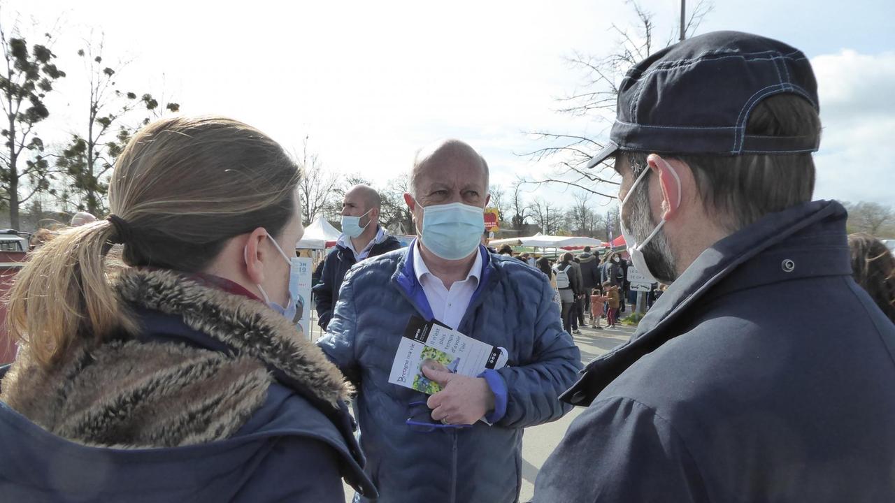 Ein Mann mit Mundschutz, blauem Anorak und Prospekten, die er vor der Brust festhält, unterhält sich mit einem Pärchen. Ein Mann mit Mundschutz, blauem Anorak und Prospekten, die er vor der Brust festhält, unterhält sich mit einem Pärchen.