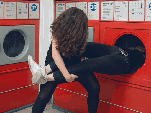 Eine Frau vor einer Waschmaschine hält die Beine eines Mannes, der mit dem Kopf und dem Oberkörper in der Waschmaschine ist.
