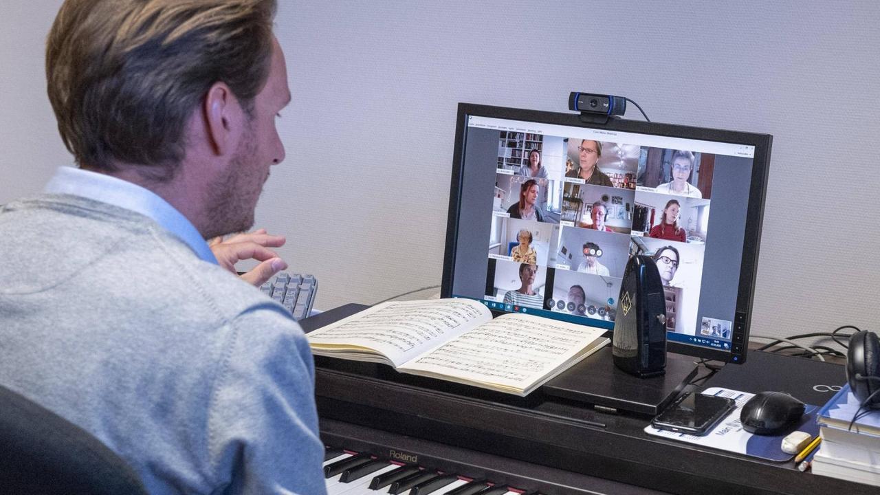 Ein Mann sitzt dirigierend an einem E-Piano, auf dem Notepult ein Bildschirm, auf dem mehrere Menschenköpfe zu sehen sind. Ein Mann sitzt dirigierend an einem E-Piano, auf dem Notepult ein Bildschirm, auf dem mehrere Menschenköpfe zu sehen sind.