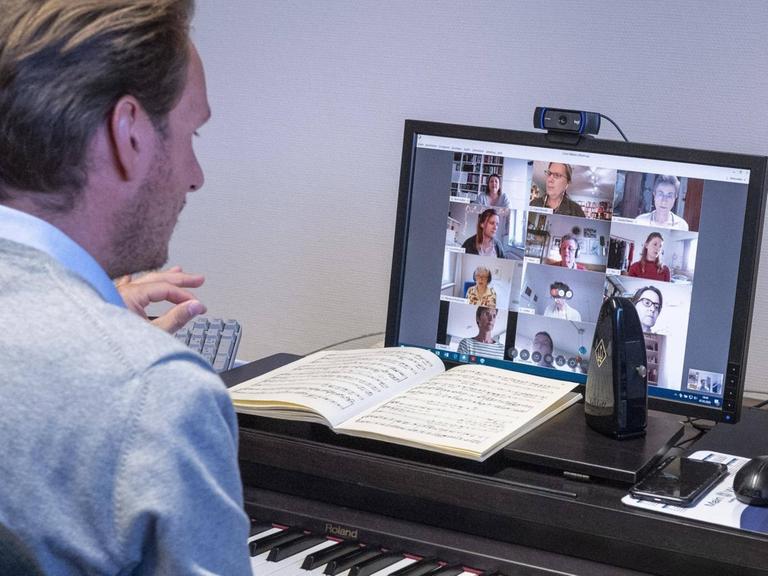 Ein Mann sitzt dirigierend an einem E-Piano, auf dem Notepult ein Bildschirm, auf dem mehrere Menschenköpfe zu sehen sind.