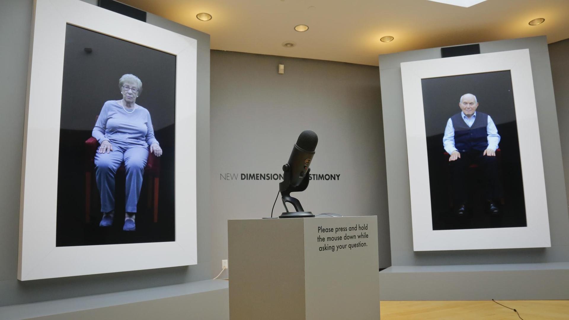In einem Aufstellungsraum befinden sich zwei Bildschirme an der Wand. Auf diesen sind zwei ältere Menschen zu sehen. In der Mitte des Raums steht ein Mikrofon. In einem Aufstellungsraum befinden sich zwei Bildschirme an der Wand. Auf diesen sind zwei ältere Menschen zu sehen. In der Mitte des Raums steht ein Mikrofon.