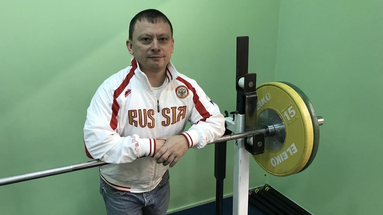 Kleiner, blasser Mann vor schweren Gewichten im Fitnesscenter in Sportjacke, auf der "Russia" steht Kleiner, blasser Mann vor schweren Gewichten im Fitnesscenter in Sportjacke, auf der "Russia" steht