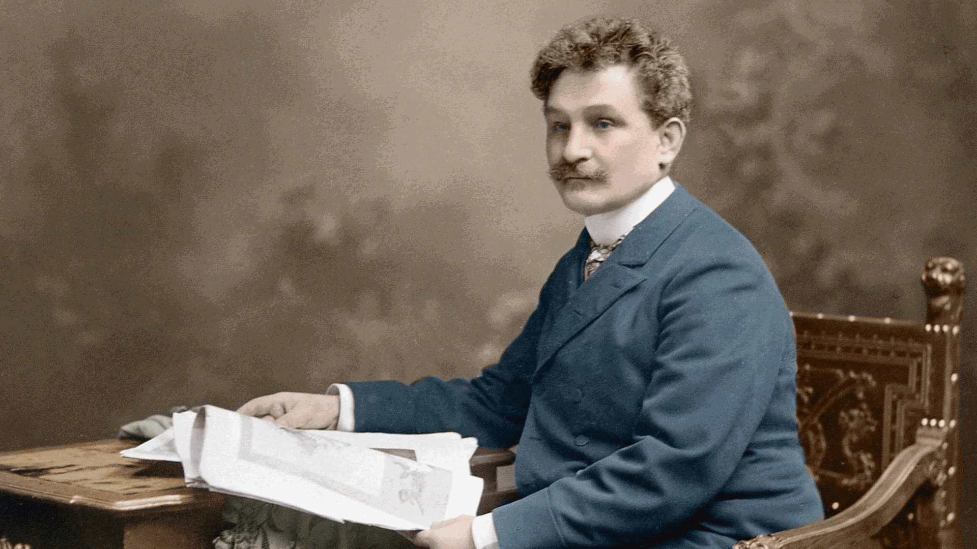 Leoš Janáček sitzt auf einer nachkolorierten Fotografie an einem Tisch und hat ein Buch aufgeschlagen. Leoš Janáček sitzt auf einer nachkolorierten Fotografie an einem Tisch und hat ein Buch aufgeschlagen.
