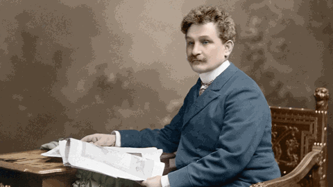 Leoš Janáček sitzt auf einer nachkolorierten Fotografie an einem Tisch und hat ein Buch aufgeschlagen.