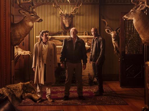 Mr Wednesday (Ian McShane, links) und Shadow Moon (Ricky Whittle, rechts) stehen mit dem Gott Vulcan (Corbin Bernsen) in einem Raum mit Hirschgeweihen und ausgestopften Tieren