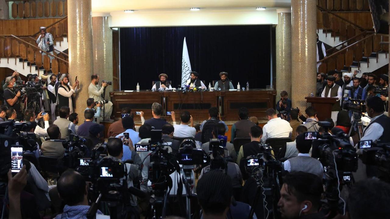 Der Taliban-Sprecher Zabihullah Mujahid sitzt mit zwei weiteren Männern am 17.08.2021 in Kabul auf einer Bühne, der Raum davor ist gefüllt mit Presseleute, die die Konferenz filmen und fotografieren. Der Taliban-Sprecher Zabihullah Mujahid sitzt mit zwei weiteren Männern am 17.08.2021 in Kabul auf einer Bühne, der Raum davor ist gefüllt mit Presseleute, die die Konferenz filmen und fotografieren.
