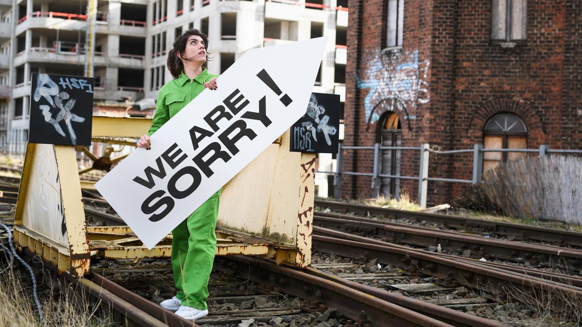 Alicia Agustín steht auf Bahngleisen und hält ein Schild mit der Aufschrift "We Are Sorry!"; im Hintergrund eine Baustelle. Alicia Agustín steht auf Bahngleisen und hält ein Schild mit der Aufschrift "We Are Sorry!"; im Hintergrund eine Baustelle.