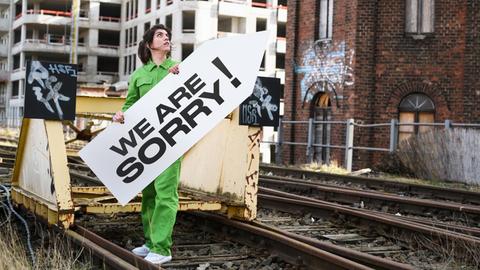 Alicia Agustín steht auf Bahngleisen und hält ein Schild mit der Aufschrift "We Are Sorry!"; im Hintergrund eine Baustelle.