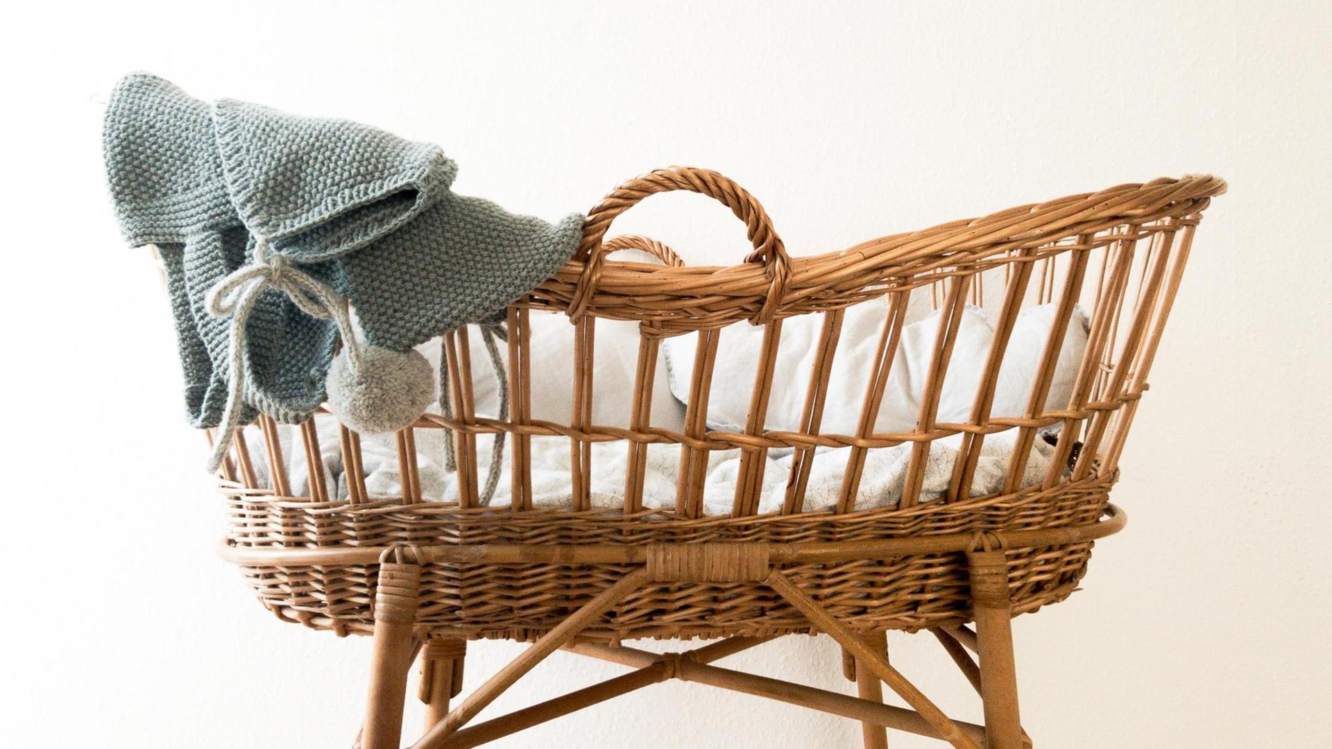 Ein leerer Stubenwagen aus Korb über dessen Rand hellblaue gestrickte Babysachen hängen. Ein leerer Stubenwagen aus Korb über dessen Rand hellblaue gestrickte Babysachen hängen.