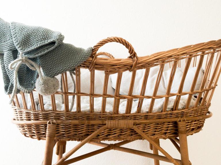 Ein leerer Stubenwagen aus Korb über dessen Rand hellblaue gestrickte Babysachen hängen.